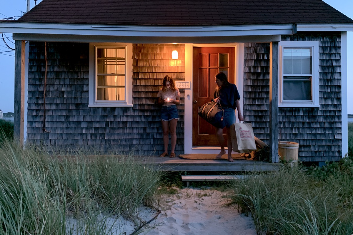 Friends at the front door of a cabin or beach house at dusk, one tapping a phone, another carrying a duffel and groceries. Warm porch light on. Candid angle.