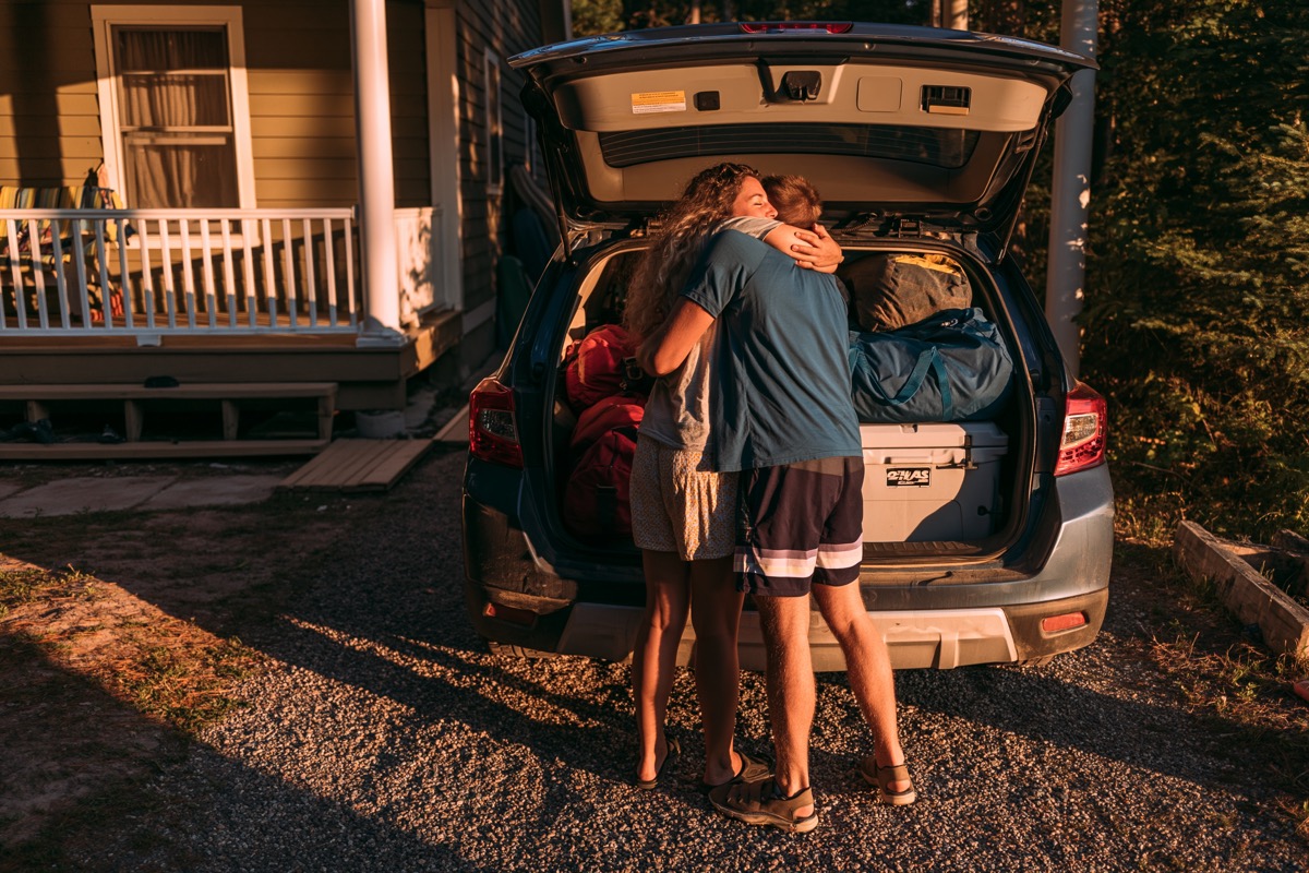 Packed car trunk, hugs in the driveway, house in the background. Bittersweet end-of-weekend warmth, golden morning light.